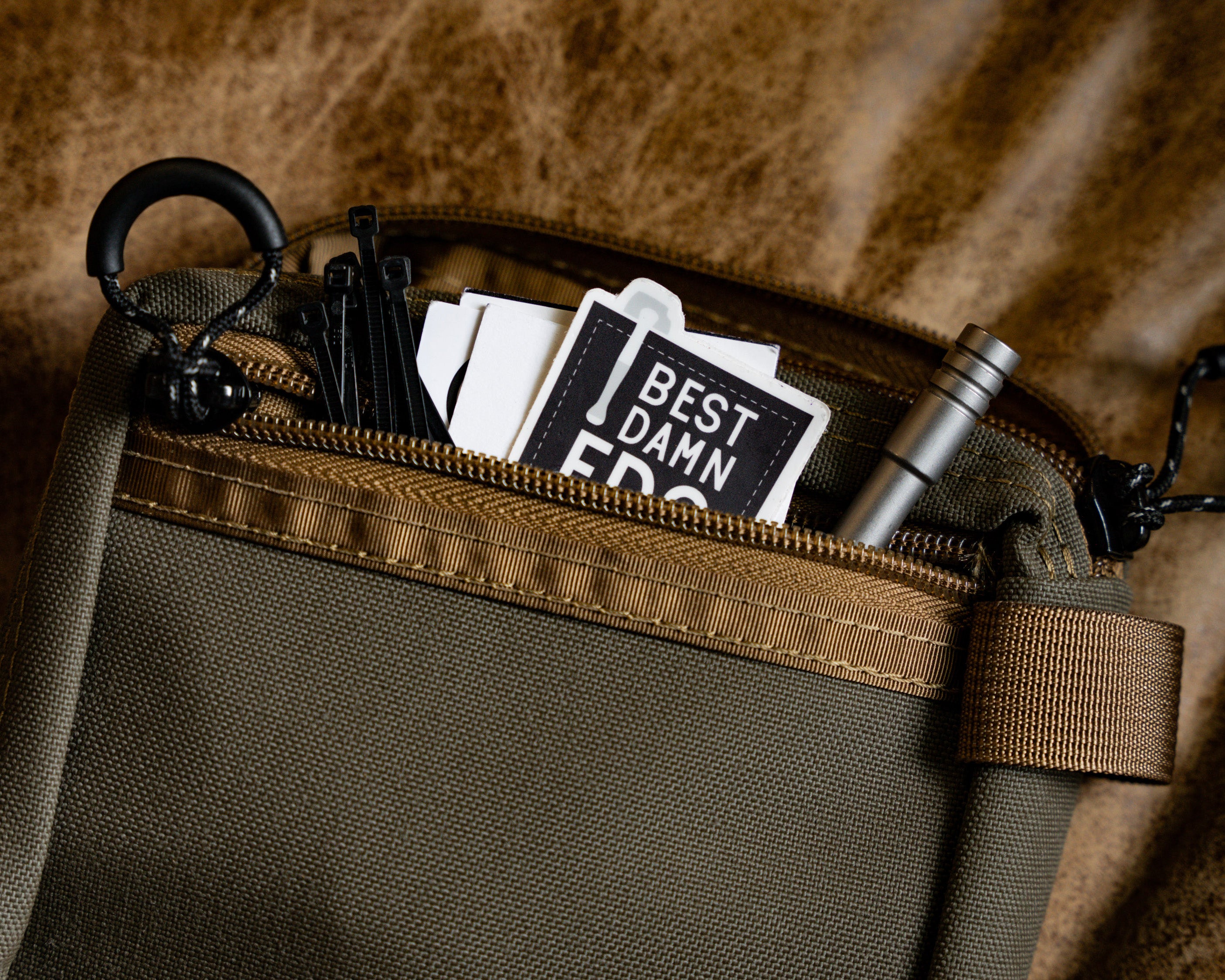 Carry Commission / Blue Ridge Overland Gear EDC Pouch Bundle view of back zippered pocket with zip ties and stickers poking out set on a leather background