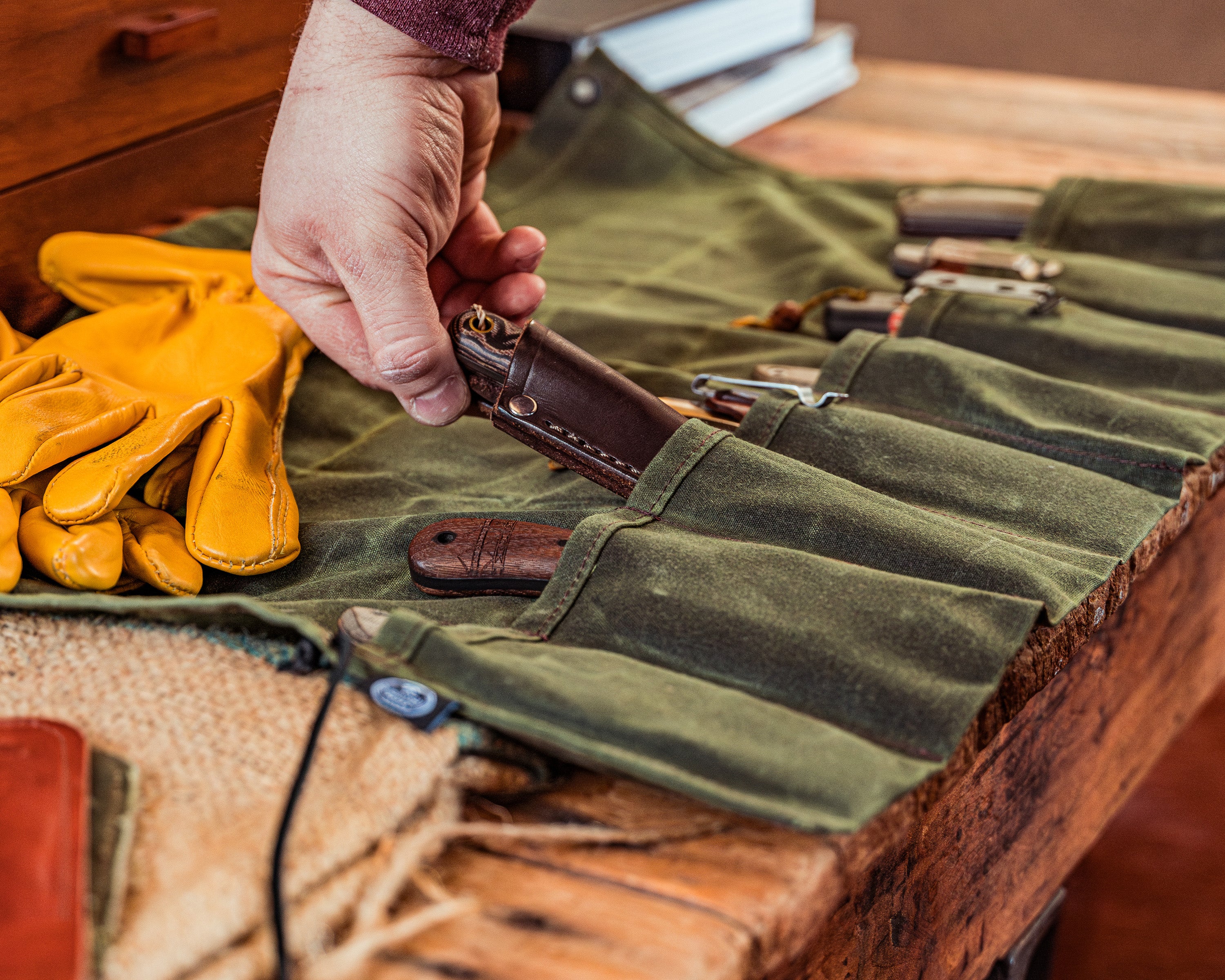 Birch Waxed Canvas Knife Roll open and filled with knives on wooden background with burlap, leather gloves and books also in the frame and a hand pulling one of the knives out of the roll