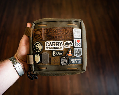 Carry Commission / Blue Ridge Overland Gear EDC Pouch Bundle front view being held by a left hand in front of a dark wood grain backgound.
