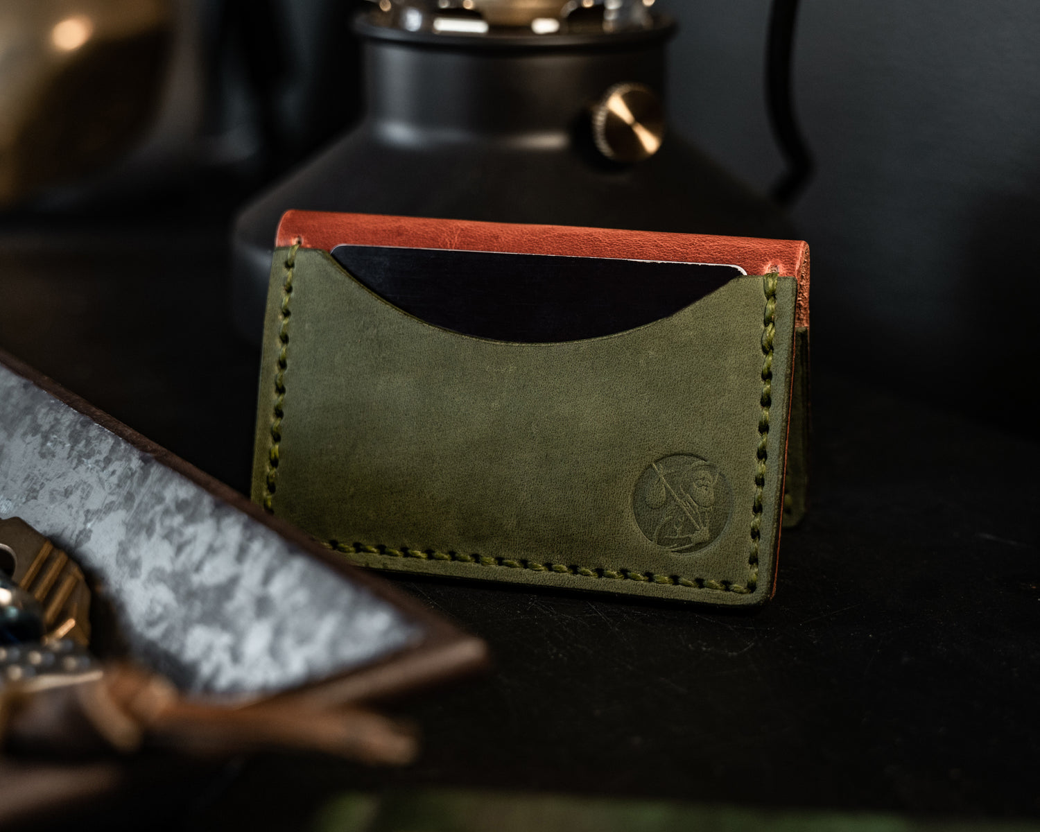 Backside view of brown and green bifold wallet in a &quot;tent&quot; shape on a black table with an edc tray in foreground and lamp in background