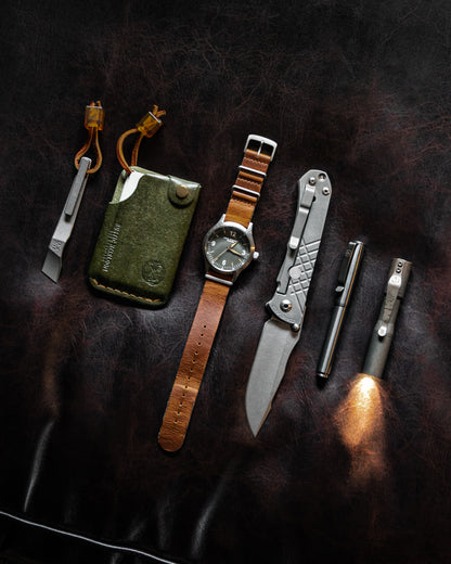 Top view of olive hitchhiker wallet, pry bar, watch, knife, pen and flashlight on a dark leather background.