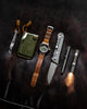 Top view of olive hitchhiker wallet, pry bar, watch, knife, pen and flashlight on a dark leather background.