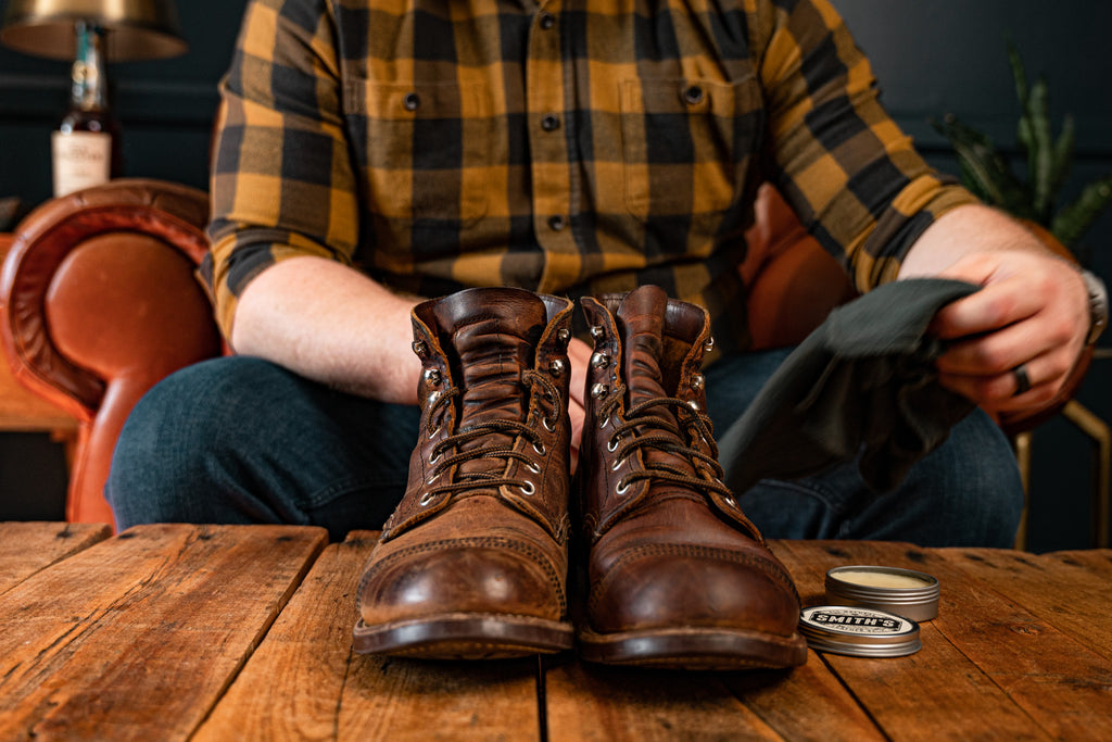 Pair of leather boots in foreground on wooden table with Smith's Leather Balm open on same table. Man in background holding Smith's Leather Balm Shop Rag.