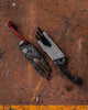 Overhead view of Best Damn EDC TPT Slide Box Cutter Tool in Black & Stonewash Topo on rust background.