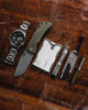 Overhead view of Best Damn EDC TPT Slide Box Cutter Tool in Stonewash Topo on wooden background pictured alongside stainless steel watch, folding knife, slim wallet, Ti pocket flashlight and mini click pen