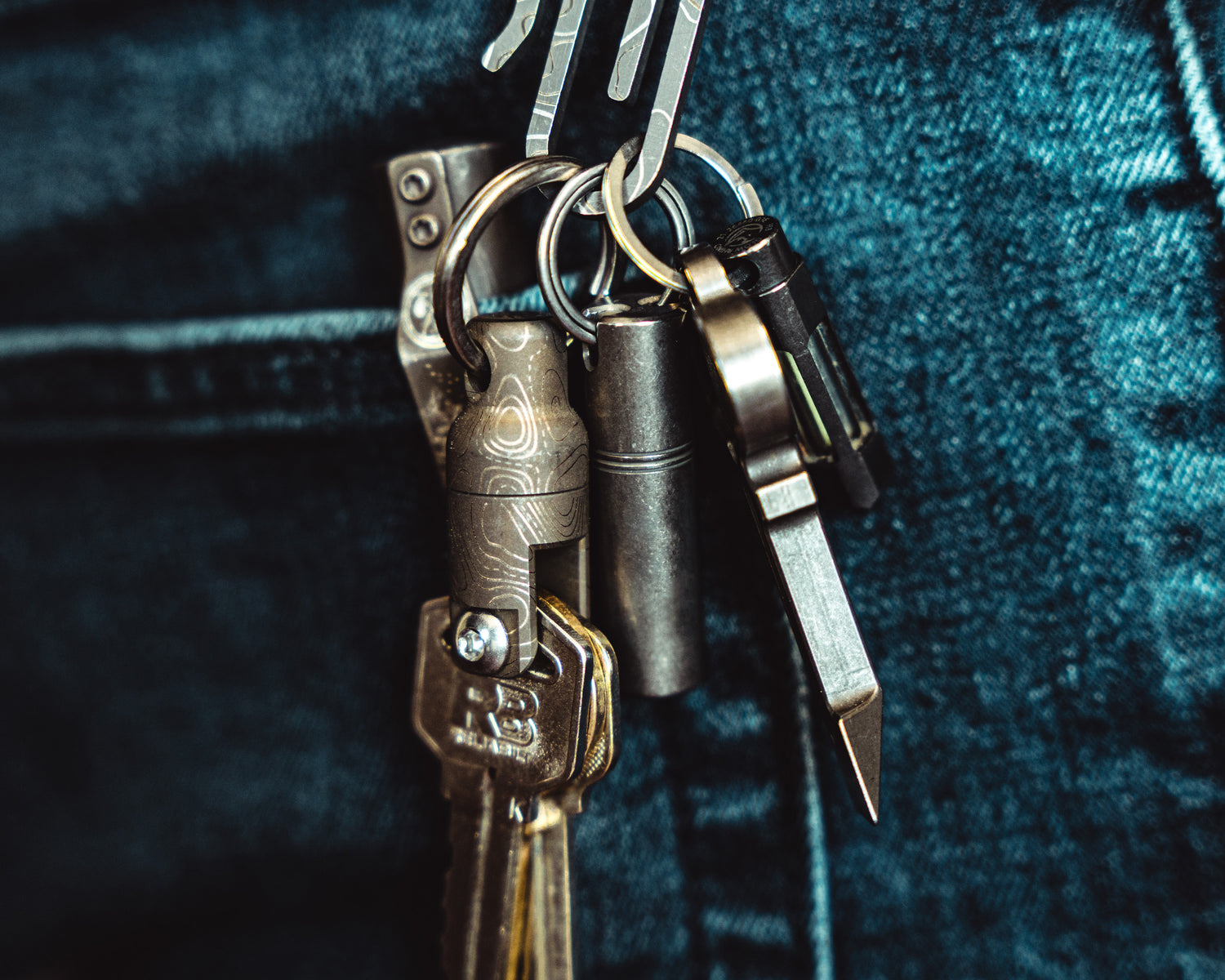 Carry Commission / Urban Carvers Keyper MQR Bronze Topo side view with keys dangling from a tough clip against a blue jean background