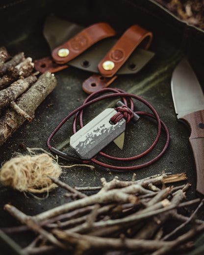 Overhead view of the Viking Spark necklace surrounded by twigs, twine, a sheath and a fixed blade on a green canvas background