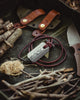 Overhead view of the Viking Spark necklace surrounded by twigs, twine, a sheath and a fixed blade on a green canvas background